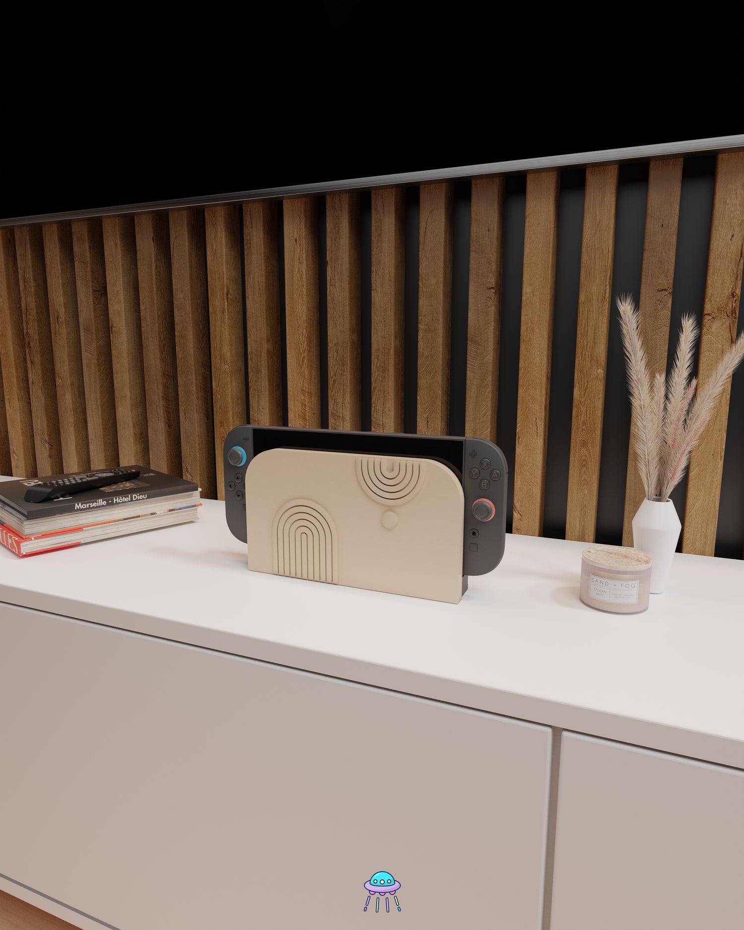 A Nintendo Switch 2 with Joy-Con controllers docked in a customised dock featuring a beige PLA filament cover and base. The cover has two embossed arch designs on the front—one larger arch at the bottom left and a smaller arch in the top right corner, alongside a small circular detail. The setup is placed on a white cabinet with a wooden panel background and decorative items nearby, including a candle, a vase with dried pampas grass, stacked books and a tv remote.