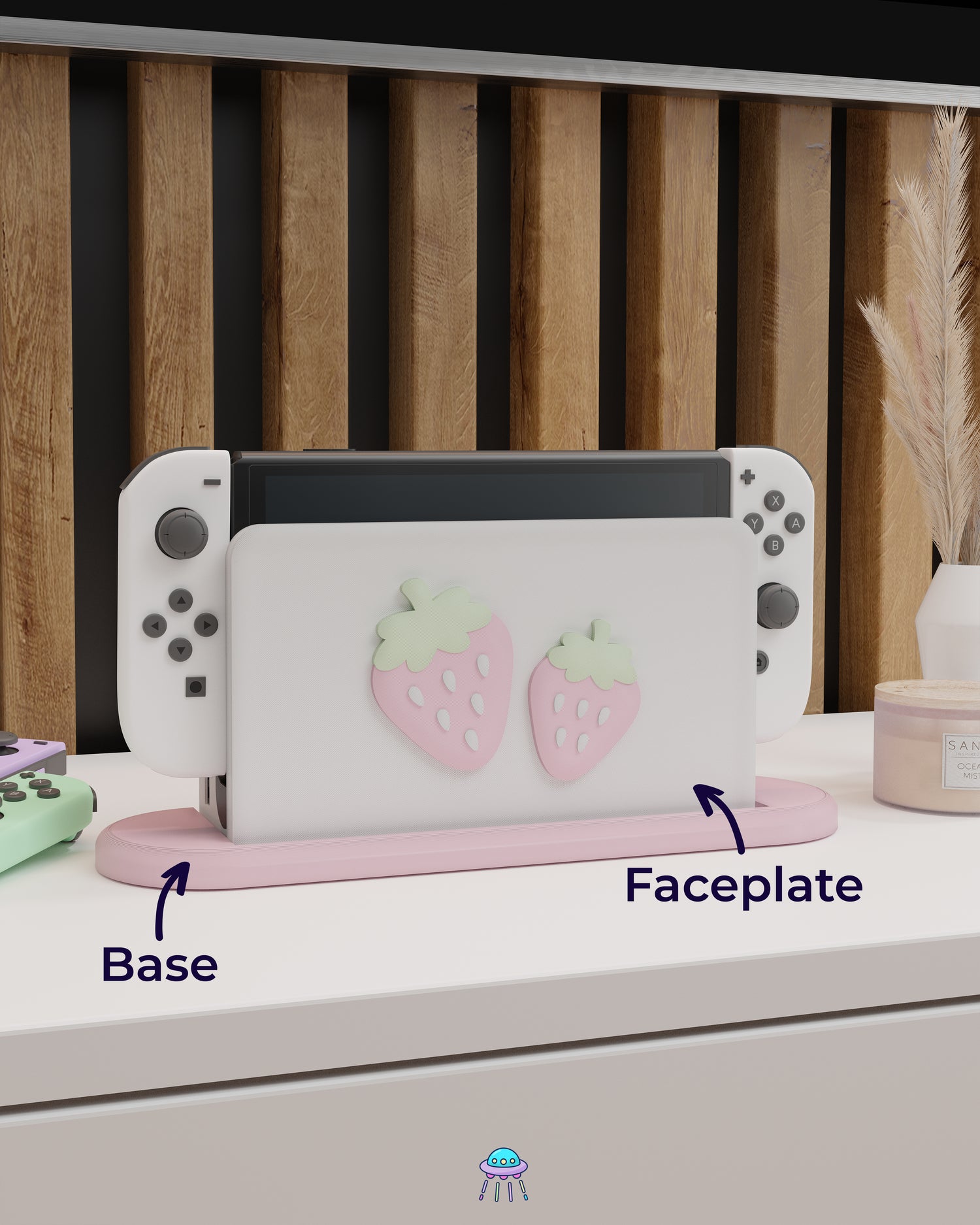 A Nintendo Switch with Joy-Con controllers docked in a customised dock featuring a white PLA filament cover and pink base. The cover has two strawberry designs on the front—one larger to the left and a smaller strawberry to the right. The setup is placed on a white cabinet with a wooden panel background and decorative items nearby, including a candle, a vase with dried pampas grass, and an additional pair of Joy-Con controllers. Arrows point to the faceplate and base.