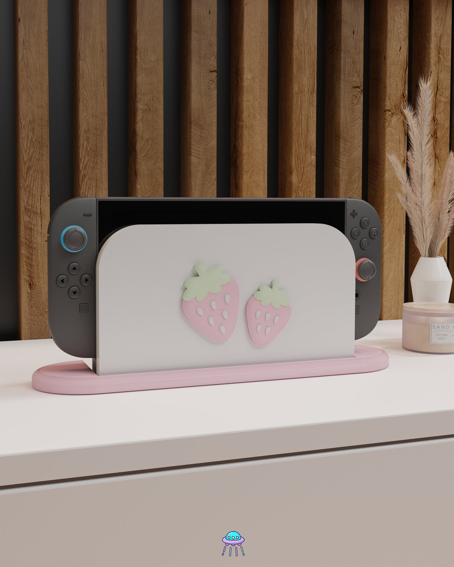 A Nintendo Switch 2 with Joy-Con controllers docked in a customised dock featuring a white PLA filament cover and pink base. The cover has two strawberry designs on the front—one larger to the left and a smaller strawberry to the right. The setup is placed on a white cabinet with a wooden panel background and decorative items nearby, including a candle and a vase with dried pampas grass.