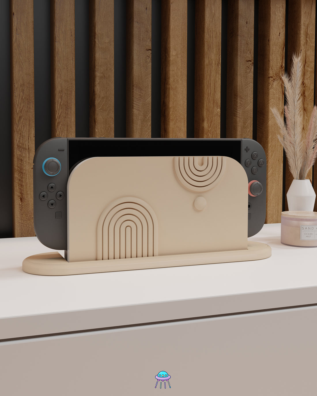 A Nintendo Switch 2 with Joy-Con controllers docked in a customised dock featuring a beige PLA filament cover and base. The cover has two embossed arch designs on the front—one larger arch at the bottom left and a smaller arch in the top right corner, alongside a small circular detail. The setup is placed on a white cabinet with a wooden panel background and decorative items nearby, including a candle and a vase with dried pampas grass.