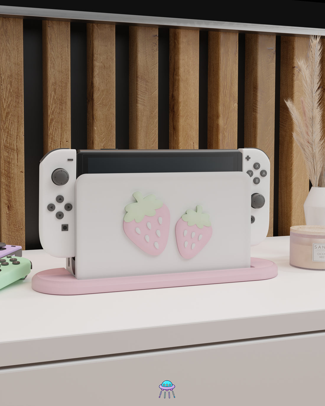A Nintendo Switch with Joy-Con controllers docked in a customised dock featuring a white PLA filament cover and pink base. The cover has two strawberry designs on the front—one larger to the left and a smaller strawberry to the right. The setup is placed on a white cabinet with a wooden panel background and decorative items nearby, including a candle and a vase with dried pampas grass.