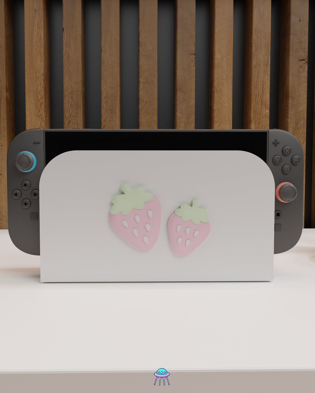 Front view of a Nintendo Switch 2 with Joy-Con controllers docked in a customised dock featuring a white PLA filament cover and pink base. The cover has two strawberry designs on the front—one larger to the left and a smaller strawberry to the right. The setup is placed on a white cabinet with a wooden panel background.